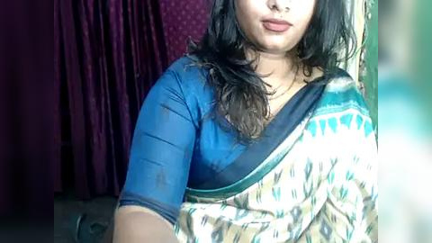 Media: Video of a South Asian woman with medium skin tone, wearing a blue blouse and a beige and green patterned sari. Background features a purple curtain and a blurred green object.