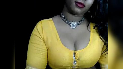 Media: Video of a woman with medium brown skin, wearing a bright yellow blouse with a low neckline, accentuating her ample cleavage, and a silver choker necklace. Dark background, focusing on her upper body.