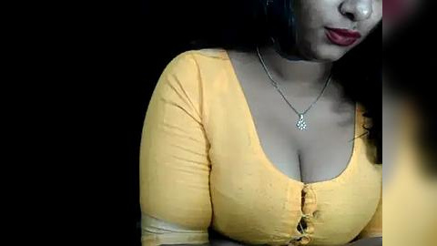 Media: A video of a woman with medium-dark skin, wearing a tight, mustard-yellow blouse that accentuates her cleavage, against a dark background. She has long, wavy black hair and wears a delicate necklace.