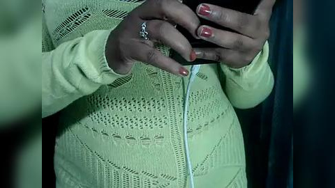 Media: Video of a dark-skinned person wearing a light green, knitted cardigan, holding a black smartphone, with red-painted nails.