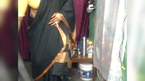 Media: Video of a person in a dark robe, holding a large, tan-colored belt, in a cluttered room with purple and green curtains, a plastic bucket, and a bottle of water.