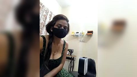 Media: Video of a woman with medium brown skin, wearing a leopard-print top, black mask, and dark hair in a bun, seated in a small, cluttered room with a mirror, shelves, and a black chair.