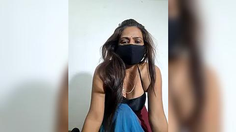 Media: Video of a young woman with long dark hair, wearing a black mask, a black top, and blue skirt, kneeling against a white wall.