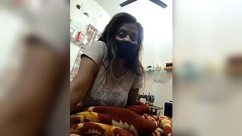 Media: Video of a South Asian woman with medium skin tone, wearing a gray T-shirt, black mask, and brown hair, sitting on a bed with a red and yellow blanket, in a cluttered bedroom with a ceiling fan.