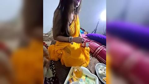 Media: Video of a woman in a bright yellow saree sitting on a rug, surrounded by colorful cushions, a plate of food, and a bottle.