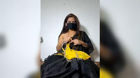 Media: Video of a woman with a dark complexion and straight, long brown hair, wearing a black mask, yellow sari, and a gold chain, seated on a wooden floor against a plain white wall.