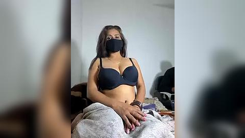 Media: Video of a woman with medium-brown skin, wearing a black bra and mask, sitting on a bed with a blanket, indoors.