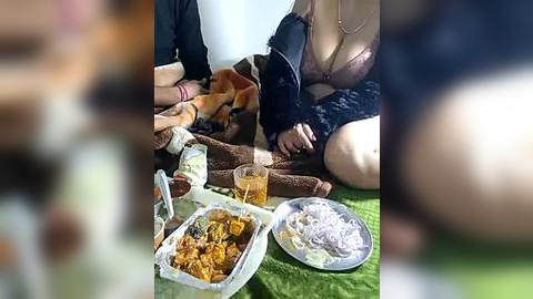 Media: Video of a woman in a black lace bra, sitting on a green blanket, eating from a tray of food, surrounded by friends, in a casual outdoor setting.