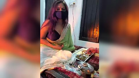 Media: Video of a South Asian woman wearing a green saree, purple blouse, and black mask, eating from a tray on a bed, surrounded by food and a red blanket.