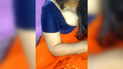 Media: Video of a woman with medium skin tone, wearing a blue velvet top and an orange sari, with a gold necklace and bracelet, seated indoors against a purple backdrop.