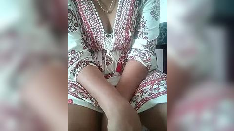 Media: A video of a woman in a white and red patterned dress with a plunging neckline, seated on a bed, legs crossed. Her fair skin is visible, and her necklace is prominent. Background is blurred.