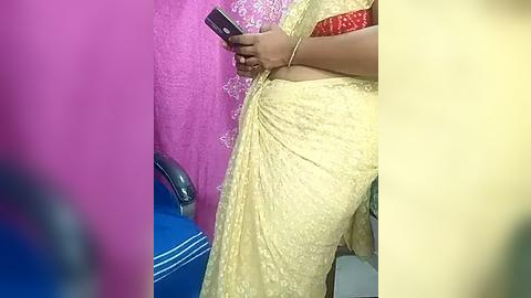 Media: Video of a woman in a yellow saree with intricate embroidery, holding a smartphone, against a backdrop of pink and blue walls.