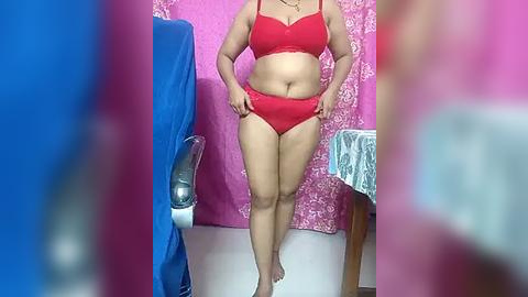 Media: Video of a plus-size woman with medium skin tone wearing red lingerie, adjusting her panties in a pink room with a blue chair and patterned curtains.