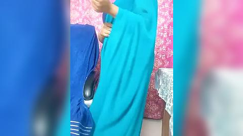 Media: Video of a person in a bright turquoise sari with blue blouse, standing in a room with pink floral wallpaper and a blue and white striped tablecloth.