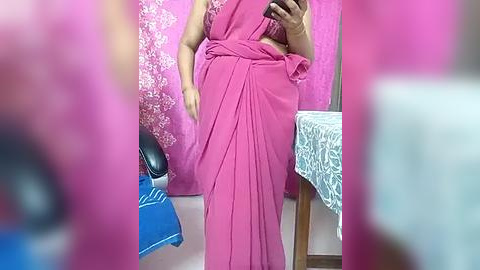 Media: Video of a person in a bright pink saree, holding a phone, in a room with pink floral curtains, blue and white patterned tablecloth, and a black chair.