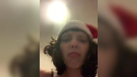 Media: Video of a woman with curly hair, wearing a Santa hat and red shirt, blurred background, dim lighting, capturing a candid moment.