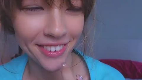 Media: A close-up video of a young woman with light brown hair, smiling broadly, wearing a turquoise shirt, and a silver necklace, set against a blurred, neutral background.