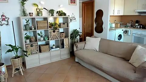 Media: Video of a cozy, minimalist living room with a beige couch, white shelving unit filled with plants and woven baskets, wooden door, guitar, and potted greenery on a tiled floor.