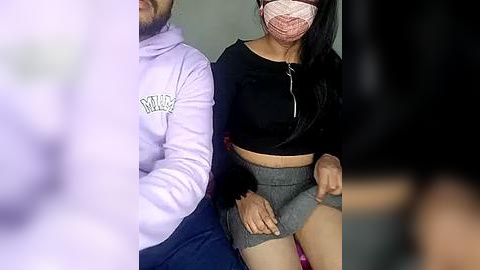 Media: A video showing a couple wearing face masks, with the man in a lavender hoodie and the woman in a black crop top and grey mini skirt, seated on a pink cushion.