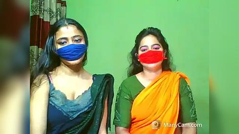 Media: Video of two women with dark hair, wearing blue and red face masks, blue and green sarees, in a green room with floral curtains.