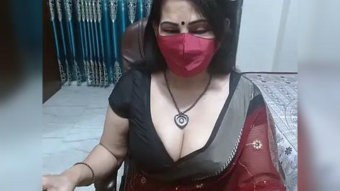 Media: Video of a South Asian woman with fair skin, wearing a red face mask, black top, and red sari, sitting on a bed in a room with blue curtains and a tiled floor.