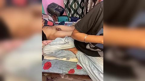 Media: Video of a person lying on a bed, partially covered by a blanket with red heart prints, in a cluttered room with hanging clothes and a pink wall.