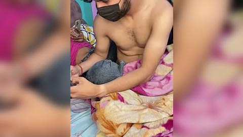 Media: Video of a shirtless South Asian man with a beard, wearing a black mask, cradling a baby on a colorful, patterned blanket. The background is blurred, emphasizing the intimate, caring moment.