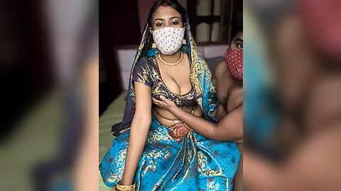 Media: Video of a South Asian woman in a blue sari with gold embroidery, wearing a face mask and gold jewelry. She is seated on a bed with a man's hand touching her breast.