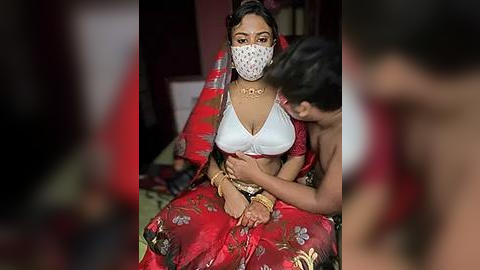 Media: Video of a woman in traditional Indian attire, wearing a face mask, with a man behind her, in a dimly lit room.