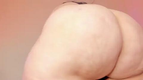 Media: A video of a nude, light-skinned person from behind, emphasizing their large, round buttocks. The background is a gradient of pink and beige.