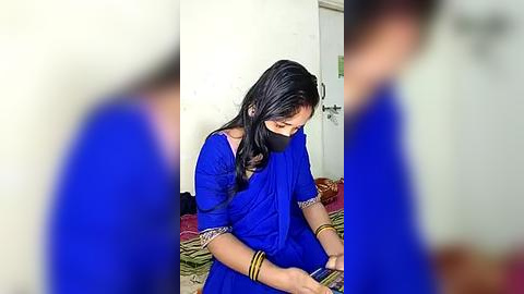 Media: Video of a young woman with long black hair, wearing a blue sari and face mask, sitting on a bed in a simple, white-walled room.