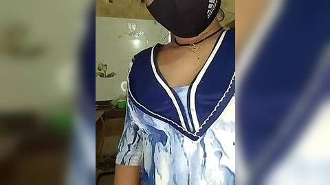 Media: Video of a person wearing a black mask, blue and white tie-dye top, and a choker necklace, in a dimly lit, cluttered room with a whiteboard and various objects.