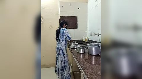 Media: Video of a woman in a blue tie-dye dress, washing dishes in a small, plain kitchen with white walls and a brown countertop.