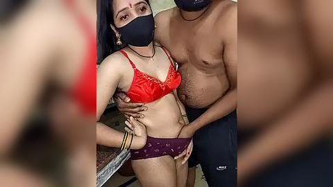 Media: Video of a South Asian transgender woman with long black hair and medium build, wearing a red lace bra and purple panties, being held by a shirtless man with a dark complexion.