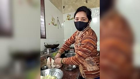 Media: A video of an Asian woman with dark hair and a black mask, wearing an orange-striped sweater, preparing food in a kitchen with white tiles and utensils on the counter.