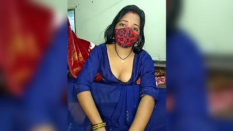 Media: Video of a woman with medium-dark skin, wearing a blue blouse, red mask, and yellow bangles, sitting in a simple room with red and white curtains.