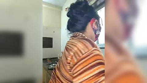 Media: Video of a woman with dark hair tied in a messy bun, wearing a striped orange and brown top, sitting in a modern kitchen with white walls, a window, and a black appliance in the background.