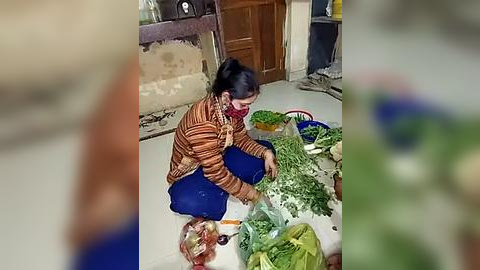 Media: Video of an Asian woman with dark hair in a ponytail, wearing a striped shirt and blue pants, squatting on a tiled floor, sorting fresh vegetables, including green leafy greens, in a kitchen with wooden cabinets and a rustic tiled counter.