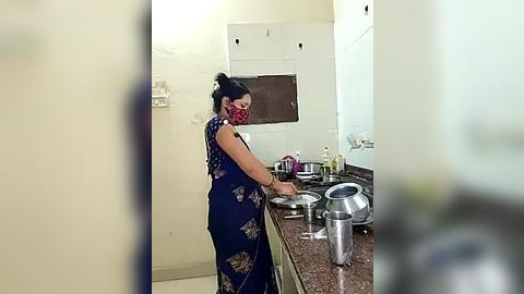 Media: A video of a woman in a dark blue floral dress, red mask, and black hair tied back, washing dishes in a small, cluttered kitchen with a stainless steel sink, brown countertops, and white walls.