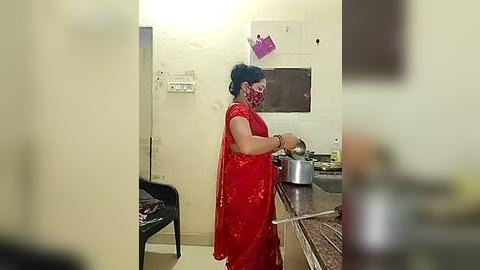 Media: A video of a woman in a red sari cooking in a small, cluttered kitchen with a stainless steel pot on a stove, a wall-mounted calendar, and a black chair in the background.