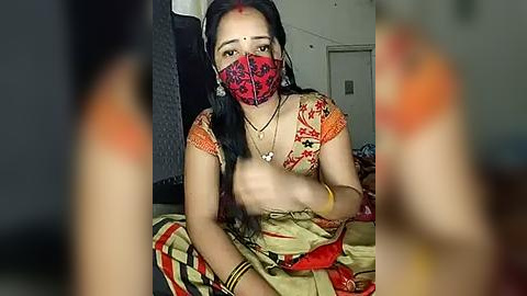 Media: A video of a young South Asian woman with long black hair, wearing a red and gold saree with a red mask, sitting indoors with a blurred background.