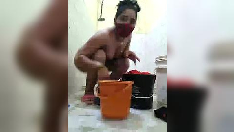 Media: Video of a woman squatting on a tiled bathroom floor, wearing a red mask, scrubbing with a sponge, surrounded by buckets and cleaning supplies.