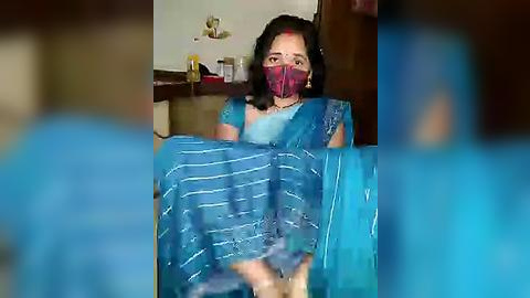 Media: A video shows a woman in a blue sari with a face mask, holding a blue sari with white stripes, in a home with wooden furniture and a shelf.