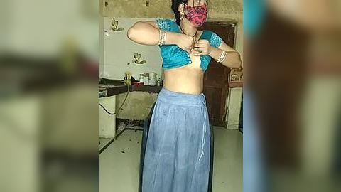 Media: Video of a woman in a kitchen, lifting her teal top, wearing a pink mask, and a blue skirt. The background shows a cluttered countertop, sink, and tiled walls.