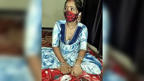 Media: Video of an Indian woman in a blue sari with red pattern, wearing a red mask, sitting on a red-patterned bed, hands tied with a red cloth, in a dimly lit room.