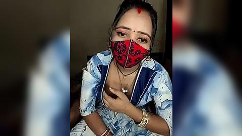 Media: Video of an Indian woman with medium skin tone, wearing a red mask and blue floral-patterned top, sitting indoors with blurred background.