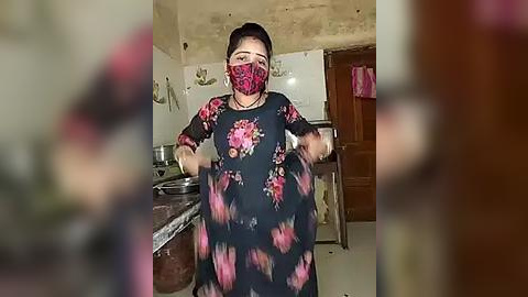 Media: Video of a woman in a dark floral dress with red and pink patterns, wearing a red mask, cooking in a simple kitchen with tiled walls and a metal sink.