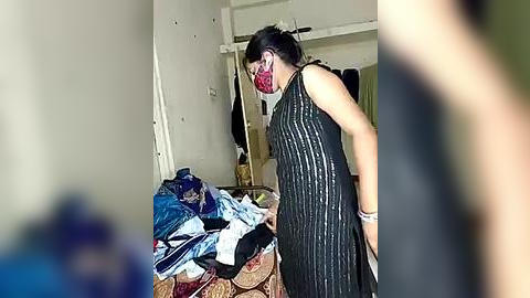 Media: A video of a woman in a black dress with vertical stripes, wearing a red face mask, standing amidst a cluttered room with clothes scattered on the floor, white walls, and a partially open door.