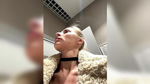 Media: Video of a woman with fair skin, light brown hair, wearing a fluffy beige jacket and black choker, looking upward in a modern, dimly lit room with white walls and a vented ceiling.