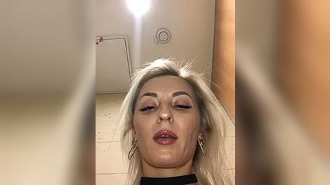 Media: Video of a young woman with long, platinum blonde hair, wearing makeup, standing in a bathroom with beige tiles and a light fixture overhead.